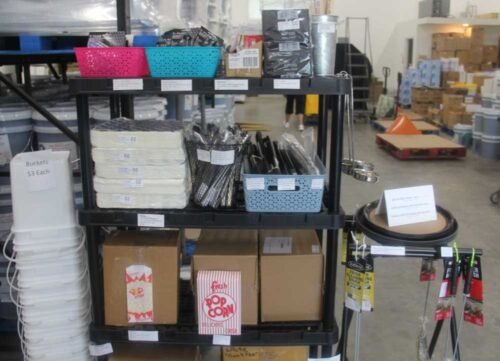 photo of a stock shelf with popcorn bags, serving ladles, and other party serving supplies with 5 gallon buckets in the background at C&C Suppliers